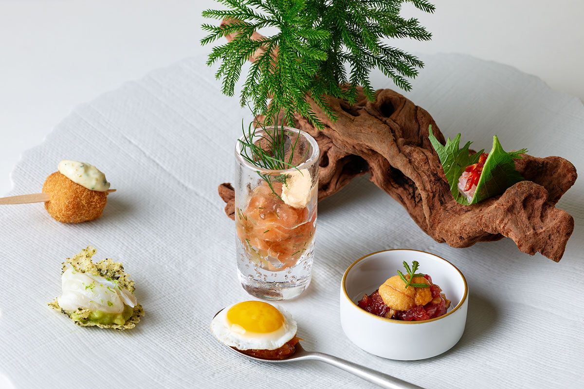 料理はコース（ランチ1万2100円～、ディナー2万4200円～）の一例。アミューズ6種。左奥から時計回りに、ピエドコションとタコのコロッケ、サーモンとフヌイユのジュレと燻製パプリカのクリームチーズ、大葉で巻いた甘エビとチョリソー、馬肉のタルタルと雲丹、いぶりがっこ 焦がし玉ねぎソース和え、ラタトゥイユとウズラ卵の目玉焼き、パルメザンチーズと青海苔のチュイルアボカドのピュレ ライムでマリネしたスズキ。