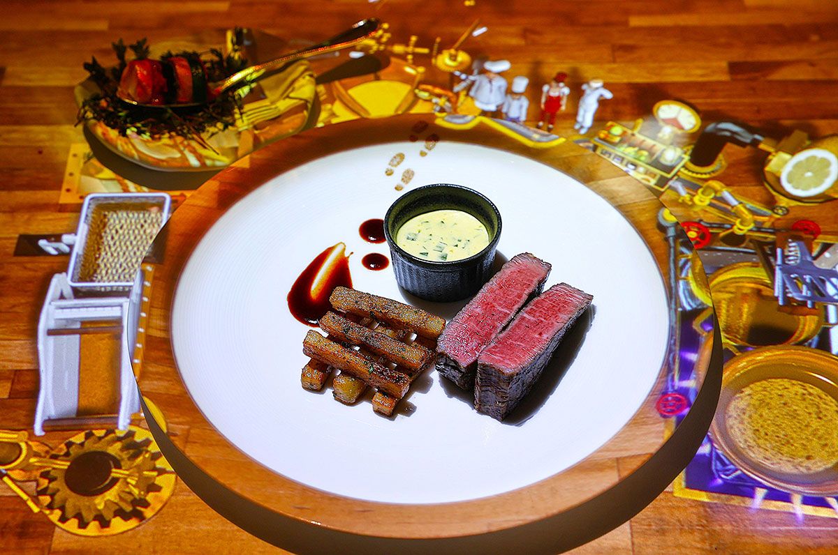 ベアルネーズソースとフレンチフライを添えた「国産牛サーロインのステーキフリット」。皿の周りにはル・プチシェフが最先端マシーンでステーキフリットを作り上げる様子が映し出されている。